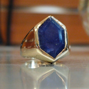 May include: A gold-toned ring with a large, deep blue gemstone in a hexagonal shape. The ring has a thick band and a polished finish. The gemstone is set in a gold bezel. The text "Silver Screen" is visible on the surface.
