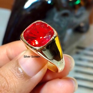 Op de afbeelding: Een goudkleurige ring met een grote, vierkant geslepen, felrode edelsteen in een gepolijste metalen band. De ring heeft een glad, reflecterend oppervlak en een opvallende, gefacetteerde rode steen. De tekst Silver Screen is zichtbaar.