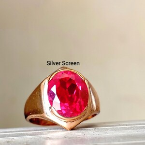Op de afbeelding: Een goudkleurige ring met een grote, ovaal geslepen, felrode edelsteen in een gladde metalen band. De tekst "Silver Screen" staat boven de ring gedrukt. Het ontwerp heeft een gepolijste afwerking met een opvallende, gefacetteerde centrale steen.