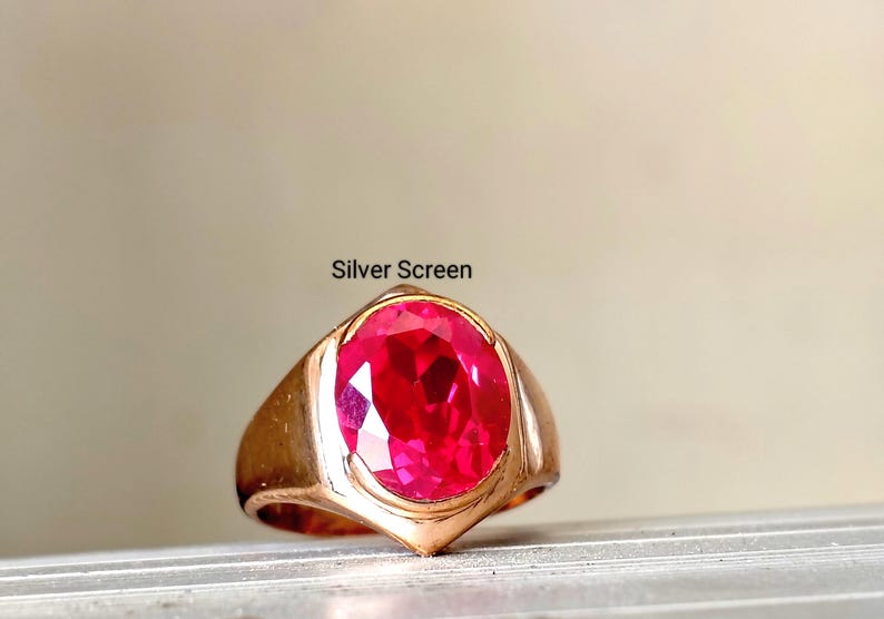 Op de afbeelding: Een goudkleurige ring met een grote, ovaal geslepen, felrode edelsteen in een gladde metalen band. De tekst "Silver Screen" staat boven de ring gedrukt. Het ontwerp heeft een gepolijste afwerking met een opvallende, gefacetteerde centrale steen.