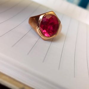 Op de afbeelding: Een goudkleurige ring met een grote, ovaal geslepen, felrode edelsteen gezet in een gladde, gepolijste metalen band. De ring heeft een opvallende geometrische vorm rond de zetting en een gefacetteerd oppervlak dat het licht reflecteert.