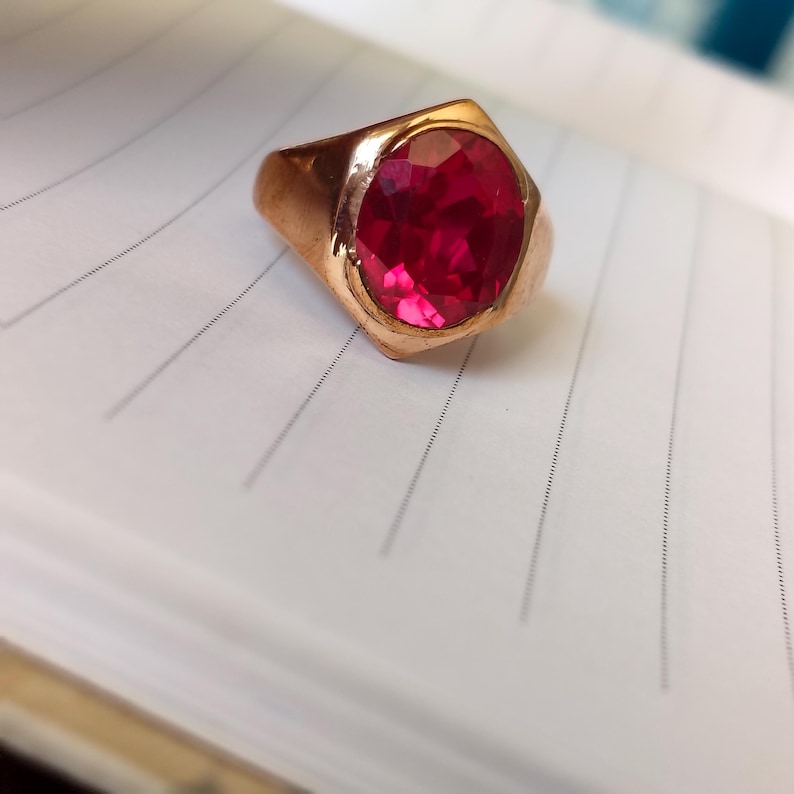 Op de afbeelding: Een goudkleurige ring met een grote, ovaal geslepen, felrode edelsteen gezet in een gladde, gepolijste metalen band. De ring heeft een opvallende geometrische vorm rond de zetting en een gefacetteerd oppervlak dat het licht reflecteert.