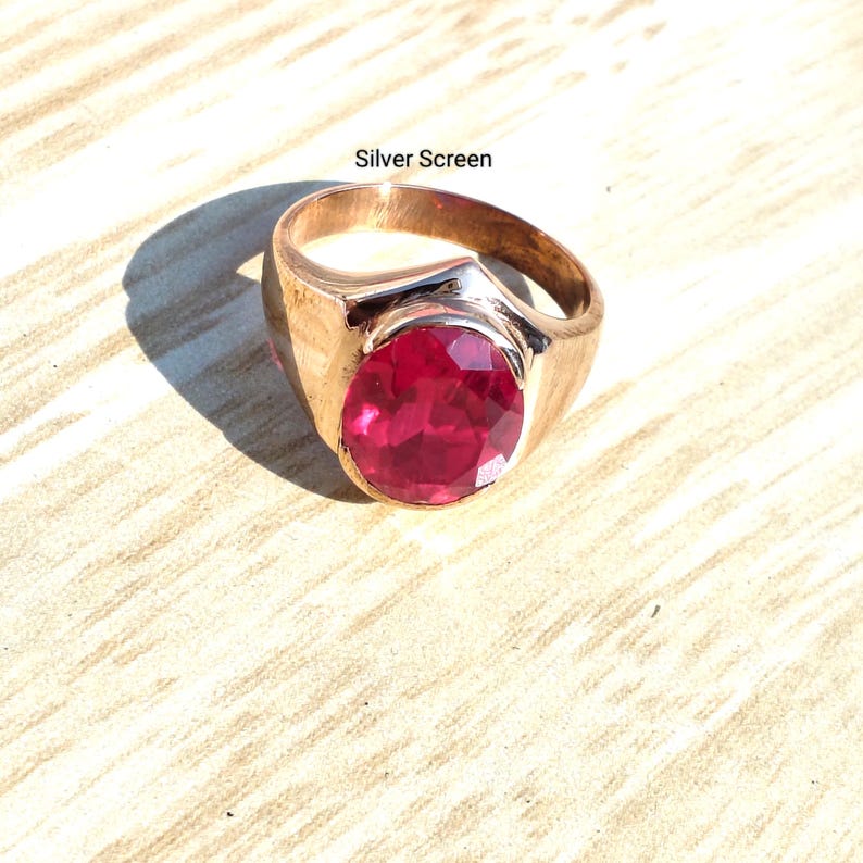 Op de afbeelding: Een goudkleurige ring met een grote, ovaal geslepen, dieprode edelsteen in een gepolijste metalen band. De tekst "Silver Screen" staat boven de ring gedrukt. Het ontwerp heeft een gladde, brede band die naar de achterkant toe smaller wordt.