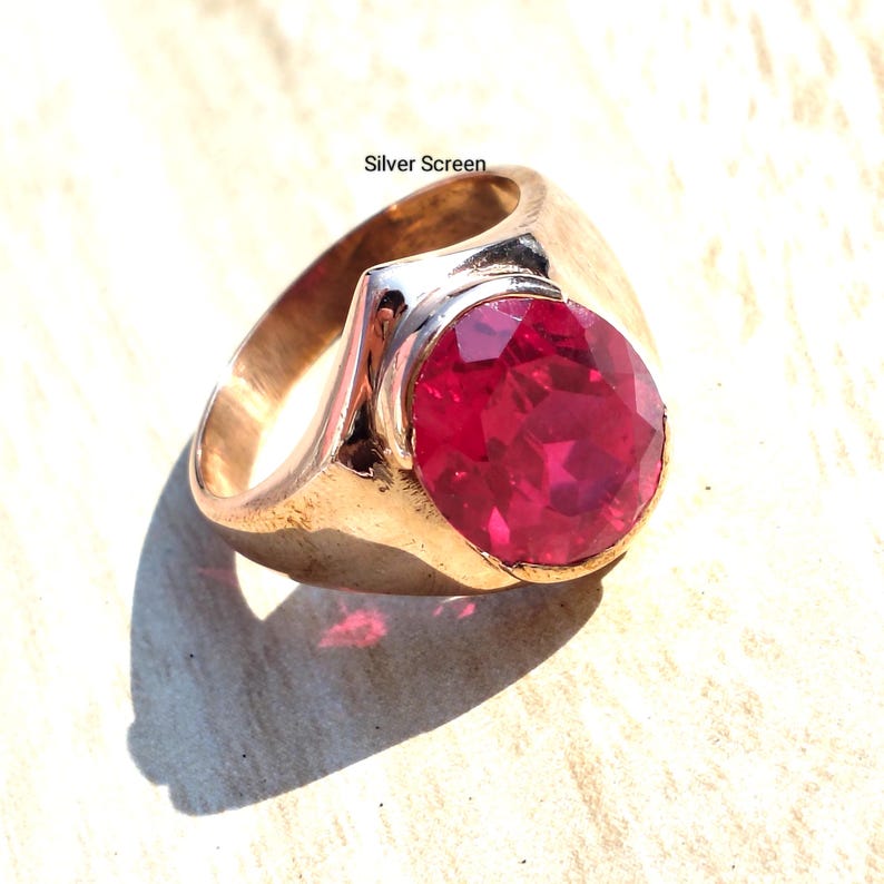 Op de afbeelding: Een goudkleurige ring met een grote, ronde, gefacetteerde dieprode edelsteen in het midden. De metalen band heeft een gladde, gepolijste afwerking. De tekst Silver Screen staat in kleine zwarte letters boven de ring op de lichte achtergrond.