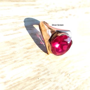 Op de afbeelding: Een goudkleurige ring met een grote, ronde, gefacetteerde, felrode edelsteen in een gepolijste metalen band. De ring is voorzien van de tekst Silver Screen en het ontwerp heeft een gladde, taps toelopende ringband die de steen ondersteunt.