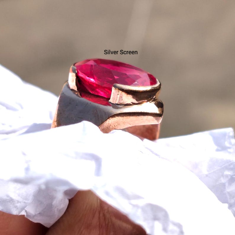 Op de afbeelding: Een ring met een grote, ovaal geslepen, felroze edelsteen in een metalen band met een koperkleurige afwerking. De tekst 'Silver Screen' staat boven de edelsteen. De ring is geplaatst tegen een verfrommeld wit materiaal.