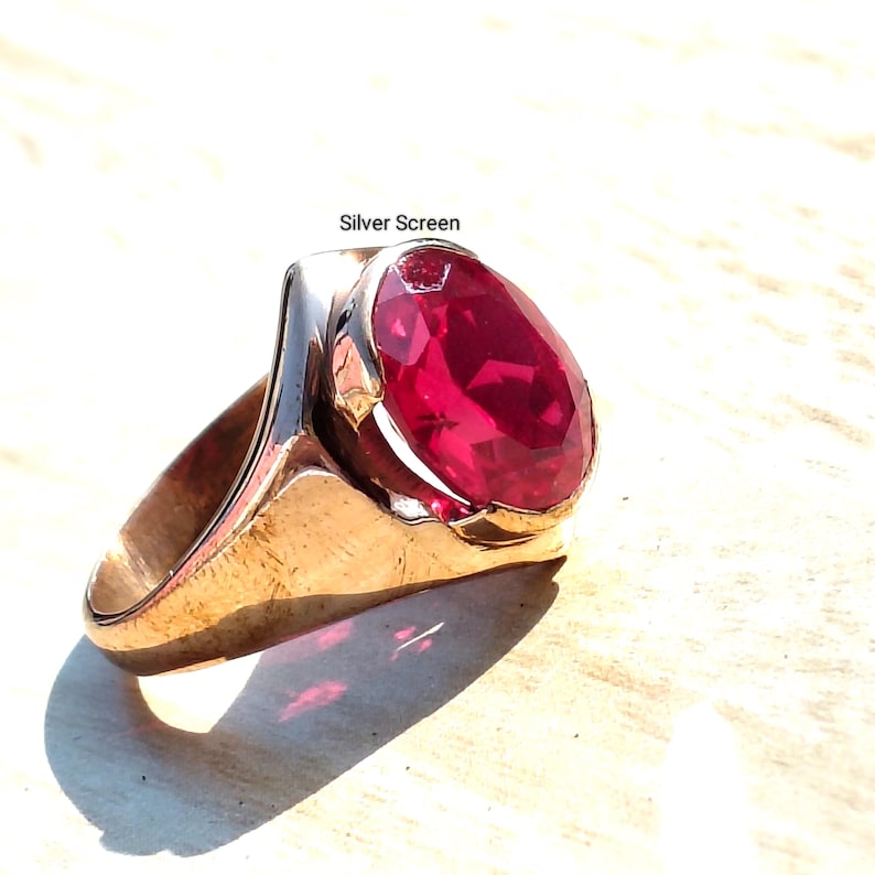 Op de afbeelding: Een goudkleurige ring met een grote, ovaal geslepen, felrode edelsteen in een gepolijste metalen band. De tekst "Silver Screen" staat boven de ring gedrukt. Het ontwerp toont een gladde, massieve ringband die taps toeloopt naar de onderkant.