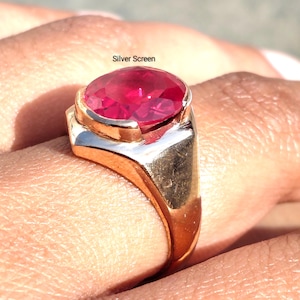 Puede incluir: Un anillo con una gran piedra preciosa roja vibrante de corte ovalado, montada en una banda de metal dorado pulido. El texto Silver Screen es visible sobre la piedra. El anillo tiene un cuerpo liso y un engaste de bisel que asegura la piedra.