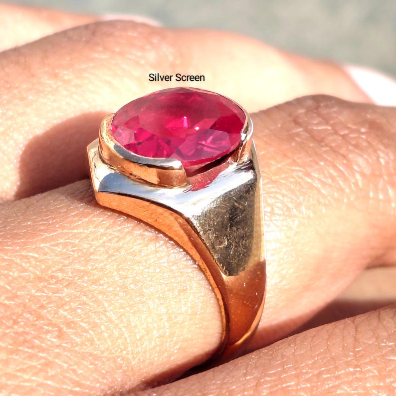 Op de afbeelding: Een ring met een grote, ovaal geslepen, levendig rode edelsteen in een gepolijste goudkleurige metalen band. De tekst Silver Screen is zichtbaar boven de edelsteen. De ring heeft een gladde, massieve schacht en een zetting die de steen vasthoudt.