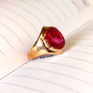 Op de afbeelding: Een goudkleurige ring met een grote, ovaal geslepen, felrode edelsteen in een gepolijste metalen zetting. De ring ligt op een gelinieerde pagina met de tekst Silver Screen in kleine zwarte letters gedrukt bij de basis van de zetting.