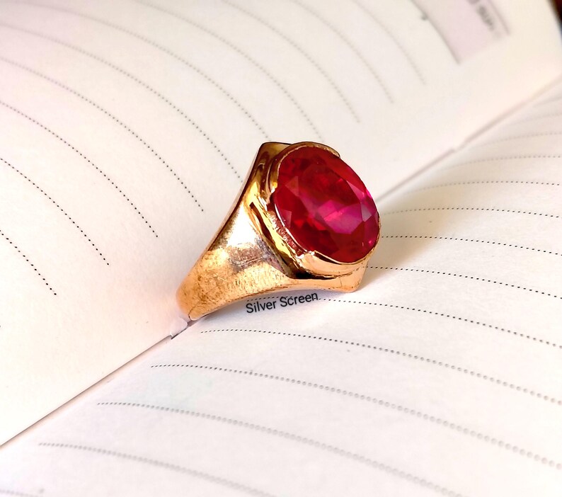 Op de afbeelding: Een goudkleurige ring met een grote, ovaal geslepen, felrode edelsteen in een gepolijste metalen zetting. De ring ligt op een gelinieerde pagina met de tekst Silver Screen in kleine zwarte letters gedrukt bij de basis van de zetting.