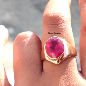 Op de afbeelding: Een ring met een grote, ovaal geslepen, felrode edelsteen in een gepolijste, goudkleurige metalen band. De band heeft een glad, breed ontwerp dat naar achteren toe smaller wordt. De tekst Silver Screen is zichtbaar boven de edelsteen.