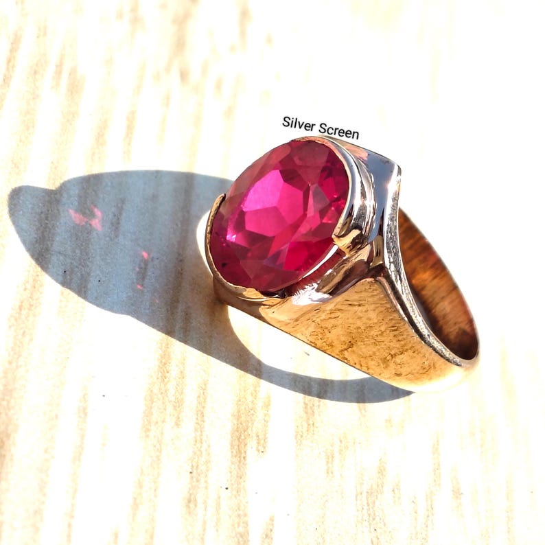 Op de afbeelding: Een goudkleurige ring met een grote, ovaal geslepen, felroze edelsteen in een gepolijste metalen band. De tekst Silver Screen staat in kleine zwarte letters boven de ring gedrukt. Het ontwerp toont een gladde, brede ringband en een gefacetteerde steen.