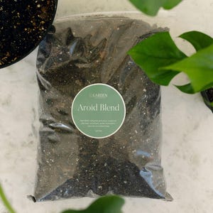 May include: A clear plastic bag filled with dark brown potting soil labeled Aroid Blend by Garden Collective. The circular green label lists ingredients including sphagnum peat moss, composted pine bark, orchid bark, perlite, and charcoal in a 1 gal bag.