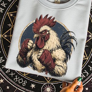 May include: A light gray crewneck sweatshirt featuring a graphic illustration of a muscular rooster with a red comb and wattle, wearing red hand wraps on its wings. The rooster is positioned inside a dark blue circle with a tan border.