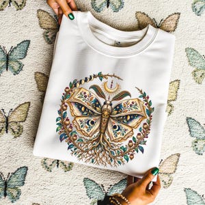 May include: A white crewneck sweatshirt featuring a central graphic of a moth with wings resembling open book pages. The moth is surrounded by a circular wreath of leaves, berries, and moon phases, with roots extending from the bottom of the moth's body.