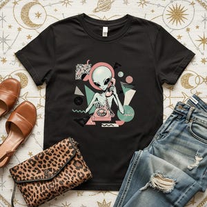 May include: A black short-sleeve t-shirt featuring a graphic print of a grey alien using a pink rotary telephone, surrounded by geometric shapes in shades of pink, teal, and black, paired with blue denim jeans, brown sandals, and a leopard print clutch.