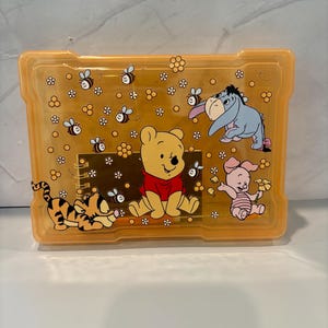 Puede incluir: Una caja de almacenamiento rectangular de plástico naranja translúcido con un diseño impreso de Winnie the Pooh, Tigger, Eeyore y Piglet. La superficie está decorada con pequeñas abejas y patrones de panal, y se ve un cuaderno de espiral dentro.