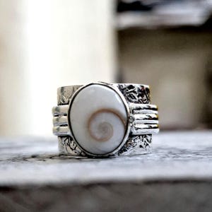 Op de afbeelding: Een zilveren ring met een centrale ovale Shiva-oogschelp met een natuurlijk bruin spiraalpatroon. De band is gemaakt van getextureerd zilvermetaal met horizontale gegroefde lijnen aan de zijkanten en een ingewikkeld, in reliëf gemaakt bloemmotief.