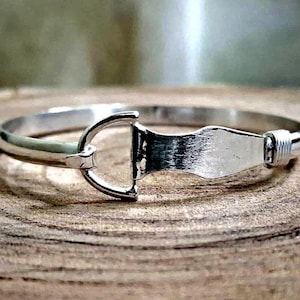 Könnte beinhalten: Ein silberfarbener Metallarmreif mit einem zentralen, vom Reitsport inspirierten Gebiss-Design. Das Armband hat eine glatte, polierte Oberfläche mit einer geschwungenen, starren Struktur und einem kleinen gewickelten Detail an einer Seite.