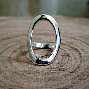 Könnte beinhalten: Ein silberner Ring mit einem ovalen Design. Der Ring hat ein dickes Band mit strukturierter Oberfläche und poliertem Finish. Die ovale Form ist in der Mitte offen und erzeugt einen einzigartigen, modernen Look. Der Ring ist vor einem Holzuntergrund platziert.