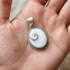 Puede incluir: Colgante ovalado con un diseño de espiral blanco y marrón, engastado en un marco de metal plateado. El colgante tiene un borde con cuentas y un lazo para una cadena.