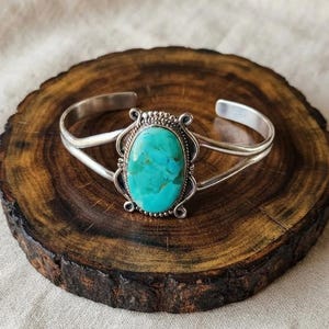 May include: A silver cuff bracelet featuring a large, oval-shaped turquoise stone set in the centre. The stone displays a vibrant blue-green colour with natural dark veining. The metal setting includes decorative beaded and twisted wire details.