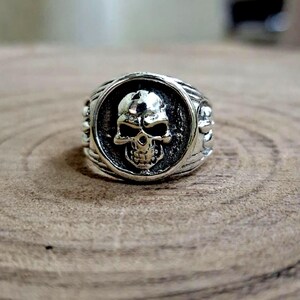 May include: Silver-toned skull ring with a detailed skull design in a circular setting. The ring has a textured band with decorative elements on the sides. The skull's features are clearly defined, creating a bold and edgy aesthetic.