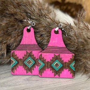 May include: A pair of pink wooden earrings with a southwestern design. The earrings are shaped like cattle tags and feature a geometric pattern in brown and teal. The earrings hang from silver-colored hooks.