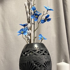 Puede incluir: Un jarrón negro, tallado intrincadamente, contiene un arreglo floral de flores azules y ramas de sauce. Una vela blanca está a la derecha. El jarrón tiene un diseño calado detallado.