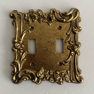 Peut inclure: Une plaque d'interrupteur double avec une finition dorée métallique et une surface texturée. Le cadre rectangulaire présente des détails floraux et des volutes en relief sur les bords, avec quatre trous de vis pour la fixation.