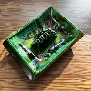 May include: A rectangular ceramic ashtray featuring a glossy glaze in shades of emerald green, lime, and dark forest green. The centre includes a raised, multi-slotted rest for cigarettes, with a smooth, reflective finish throughout the piece.