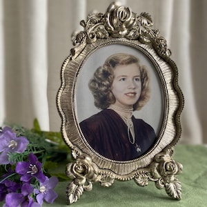 Peut inclure: Un cadre photo ovale doré avec un portrait de femme. Le cadre est orné de motifs floraux en haut et en bas. La femme porte une robe sombre et un collier de perles. Le cadre est posé sur une surface verte.