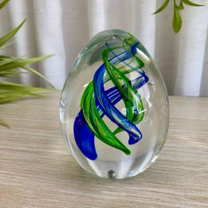 Peut inclure: Un presse-papier en verre transparent en forme d'œuf, présentant des rubans internes tourbillonnants de verre bleu vif et vert éclatant. L'objet en verre massif possède une surface lisse et polie avec de petites bulles d'air emprisonnées.