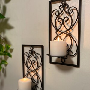 May include: Two rectangular black metal wall sconces featuring an intricate scrollwork design within the frame. Each sconce includes a circular platform holding a white pillar candle. The metal frames are mounted vertically on a flat wall surface.