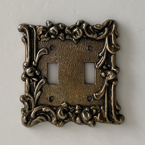 May include: A double toggle light switch plate with a metallic bronze finish. The rectangular plate features an ornate, raised relief border with scrolling leaf and floral motifs surrounding a textured, dark bronze central panel with two cutouts.