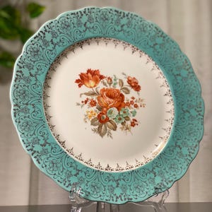 May include: A vintage decorative plate with a scalloped edge. The plate features a turquoise border with a floral design and a central floral bouquet in shades of orange, red, and yellow. The plate is displayed on a clear stand.