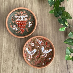 Peut inclure: Deux petites assiettes rondes en céramique avec des bords en terre cuite. L'une présente un motif de raisin rouge sur fond vert. L'autre a un motif d'oiseaux blancs et d'éléments floraux rouges sur fond marron.