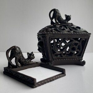 May include: A dark brown resin box with a lid featuring a sculpted cat figure and intricate openwork scroll patterns. Beside it sits a matching square tray with a low border and a sculpted cat figure, containing a stack of plain white paper sheets.