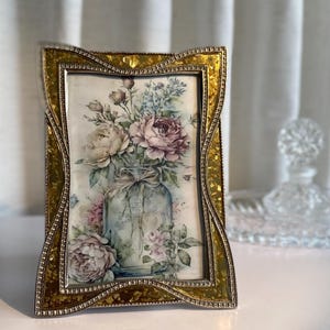 Peut inclure: Cadre photo décoratif avec une bordure dorée de style mosaïque. Le cadre contient une impression florale avec des pivoines roses et blanches dans un bocal en verre. Le cadre est rectangulaire avec un design incurvé.