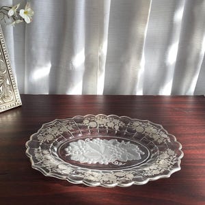 Peut inclure: Plat à servir ovale en verre transparent avec un bord festonné. Le plat présente un motif de raisin givré au centre et des motifs floraux sur le bord. Le plat est présenté sur une surface en bois foncé.