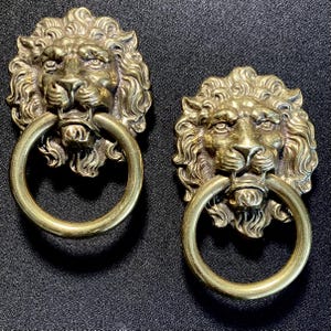 May include: Two antique brass lion head door knockers. Each knocker features a detailed lion's face with a ring in its mouth. The brass has a warm, aged patina. The knockers are mounted on a dark background.