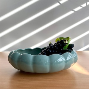 May include: A shallow, scalloped ceramic bowl in a soft sage green glaze. The bowl has a rounded, fluted edge and a smooth, glossy finish. Inside the bowl sits a cluster of dark purple grapes with green stems and leaves, creating a contrast.