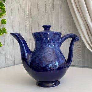 Peut inclure: Une théière en céramique avec un vernis bleu profond présentant des reflets marbrés plus clairs en bleu et blanc. La théière possède un bec verseur incurvé, une anse arrondie avec un détail enroulé et un couvercle assorti avec un bouton.