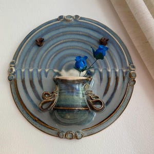 Peut inclure: Un vase mural en céramique avec un glaçage bleu et marron, présentant un design circulaire avec un petit vase pour les fleurs. Deux fleurs bleues sont placées dans le vase. Le vase a des poignées décoratives.