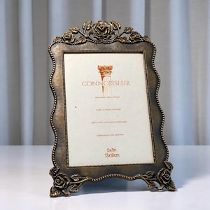 Op de afbeelding: Een antiek bronskleurige fotolijst met een rechthoekige vorm en golvende randen. De lijst is voorzien van reliëfmotieven van rozen en bladeren aan de boven- en onderkant. De inleg toont de tekst CONNOISSEUR en de afmetingen 13x18cm.