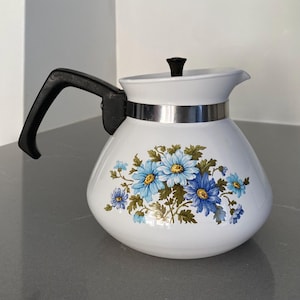 Puede incluir: Una tetera blanca con un estampado floral de margaritas azules y hojas verdes. La tetera tiene un asa negra, un pomo de tapa negro y una banda metálica de color plateado que rodea la parte superior del cuerpo debajo de la tapa.