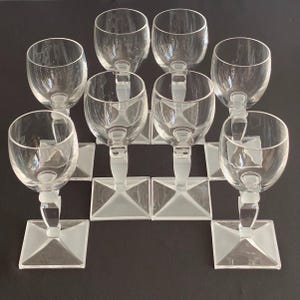 Peut inclure: Un ensemble de huit verres à pied en verre transparent disposés en grille. Chaque verre présente un bol arrondi, une tige courte avec un accent en verre dépoli et une base carrée. La structure en verre transparent offre un aspect uniforme.