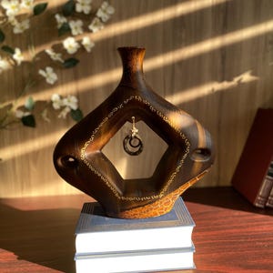 Peut inclure: Un vase en bois artisanal avec un design géométrique unique. Le vase est brun foncé avec des accents brun clair et des détails dorés. Une paire de boucles d'oreilles créoles est suspendue au centre. Le vase est posé sur une pile de livres.