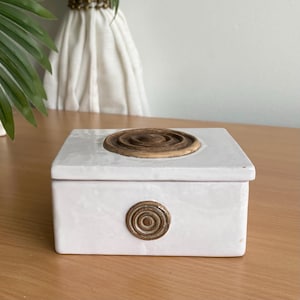 Peut inclure: Une boîte rectangulaire en céramique blanche avec une finition lisse et brillante. Le couvercle présente un motif circulaire marron en relief au centre avec des anneaux concentriques. Un emblème circulaire marron plus petit est centré sur la face avant.